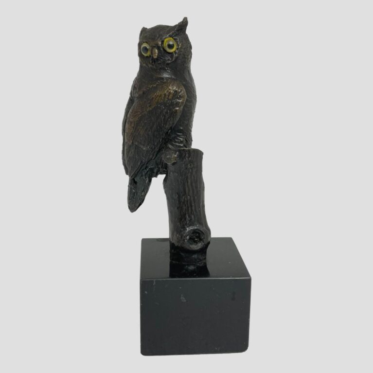 Main photo of vintage small owl bronze. The owl has his head turned to the camera so its yellow glass eyes are looking at you but its breast is facing right with tail hanging down on the left. The branch that it is perched on is not visible from this angle but the trunk can be seen. The wonderful thing about this piece is that the owl is doing what an owl does - turning its head to impossible degrees!