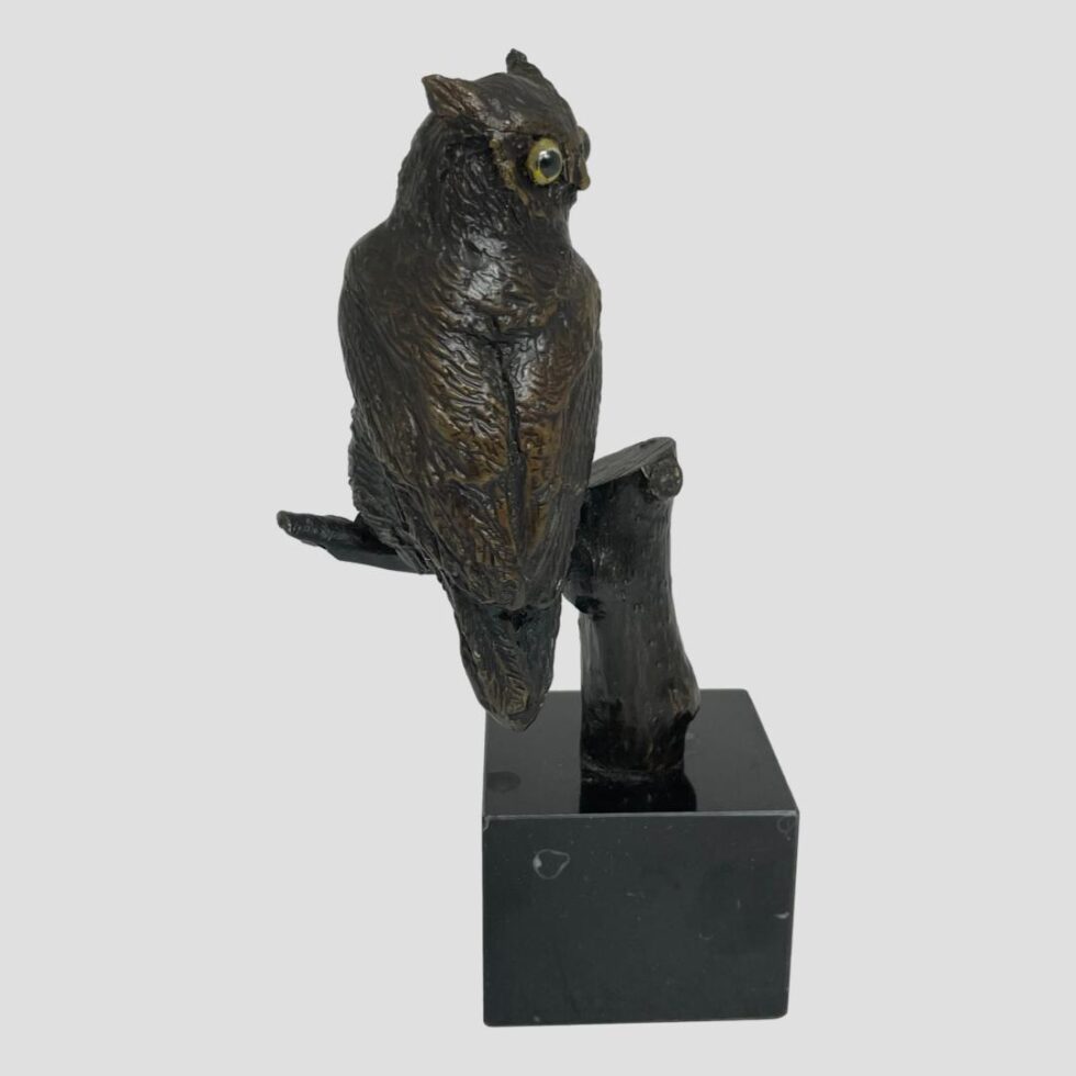 Photo of vintage small bronze owl this time seen with the back of the bird on display and the owls head facing to the right. One yellow glass eye is visible. As the owl is perched on a tree trunk, it is sticking out slightly to the left.