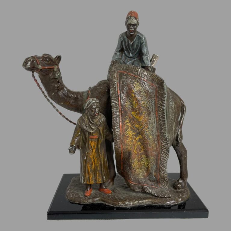 Main photo of spelter rug merchants displayed with the 'selling' side of camel on display namely, the rug is hanging rolled out from the camels hump. It is held up by a merchant in a red fez hat. On the ground is a merchant in a turban hawking the rug to passers by. A lot of the paint has worn off but it is still possible to make out some colours especially the red and orange in the rug and the red hat, slippers, camels harness and light blue tunic of man on camels back. This is a lovely antique piece in original condition.