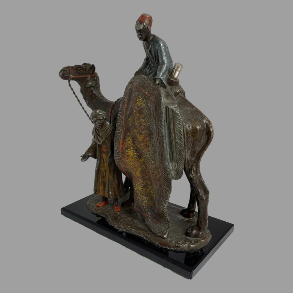 Photo of rug merchants taken from back left side of camel. The strike area is visible between the camel and the rug. The seated merchant is looking downward to the bottom left corner and the camel is looking to the top left.