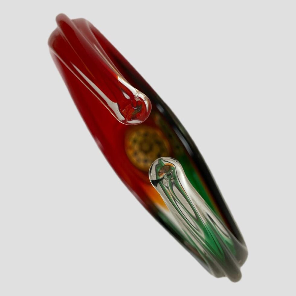 Photo of Murano basket displayed diagonally on the page. The red handle side is on the top left and green in bottom right. The circle of the millefiori base is just visible. The handles curve over the top just short of touching by an inch and have nice smooth rounded ends.
