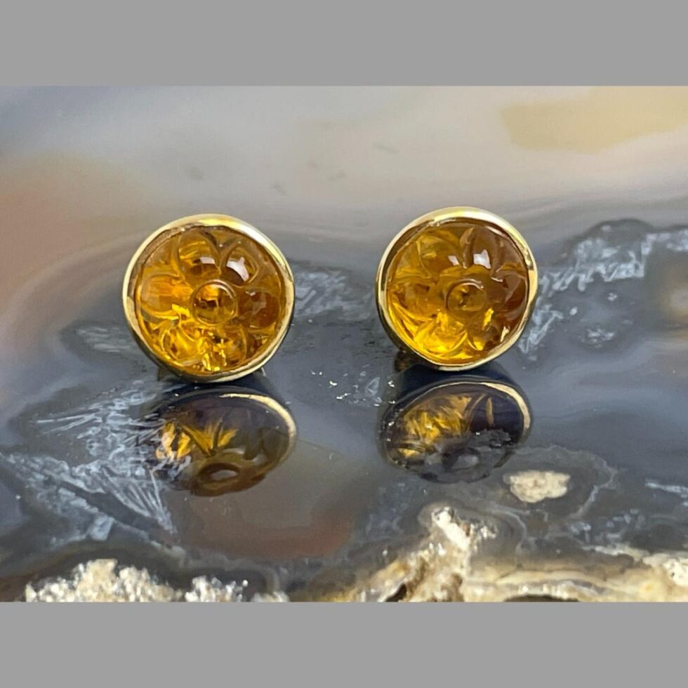 A close up photo of the carved citrine gold studs displayed outside of their box on a slice of natural agate. Both the earrings are facing forward and the carved flower design is clearly visible. The flowers look like daisies. The citrine is a lovely orange colour which compliments the 18 karat gold colour beautifully.