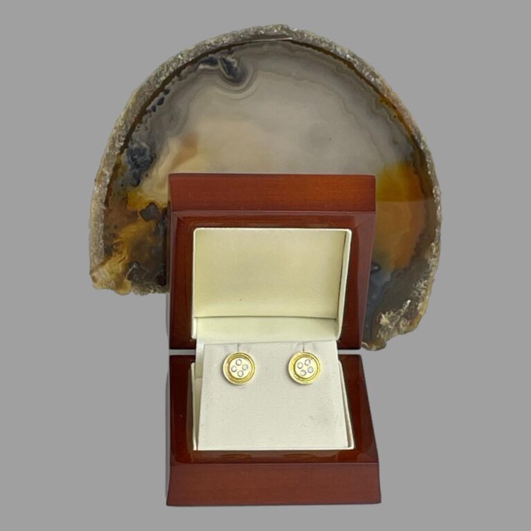 Main photo of gold diamond 'button' earrings displayed inside their box with the earring fronts forward facing. The button shape of the earrings is clear. The lacquered wooden box is a reddish brown hue and there is a slice of natural agate displayed upright behind the open lid.