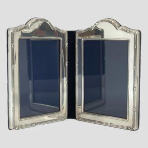 Main photo of silver double photo frame seen from an eye level with the 2 frames slightly inward facing to prop the frame open. The shape of the frames are a rectangular with a raised bump area to the top of each. The central folding area is blue velvet and this is the area with wear as it is most flexible. The design on the silver is what may be considered classical. 3 lines around the photo space area and 3 again on outer area except the outer lines have crosses spaced out all around. The area for the photographs is of a generous size