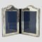 Main photo of silver double photo frame seen from an eye level with the 2 frames slightly inward facing to prop the frame open. The shape of the frames are a rectangular with a raised bump area to the top of each. The central folding area is blue velvet and this is the area with wear as it is most flexible. The design on the silver is what may be considered classical. 3 lines around the photo space area and 3 again on outer area except the outer lines have crosses spaced out all around. The area for the photographs is of a generous size