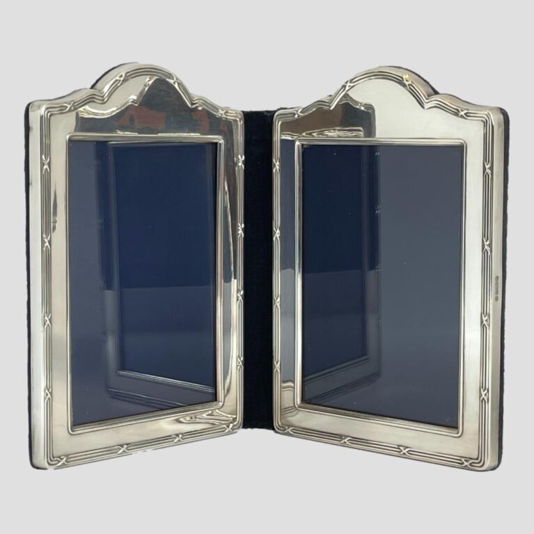 Main photo of silver double photo frame seen from an eye level with the 2 frames slightly inward facing to prop the frame open. The shape of the frames are a rectangular with a raised bump area to the top of each. The central folding area is blue velvet and this is the area with wear as it is most flexible. The design on the silver is what may be considered classical. 3 lines around the photo space area and 3 again on outer area except the outer lines have crosses spaced out all around. The area for the photographs is of a generous size