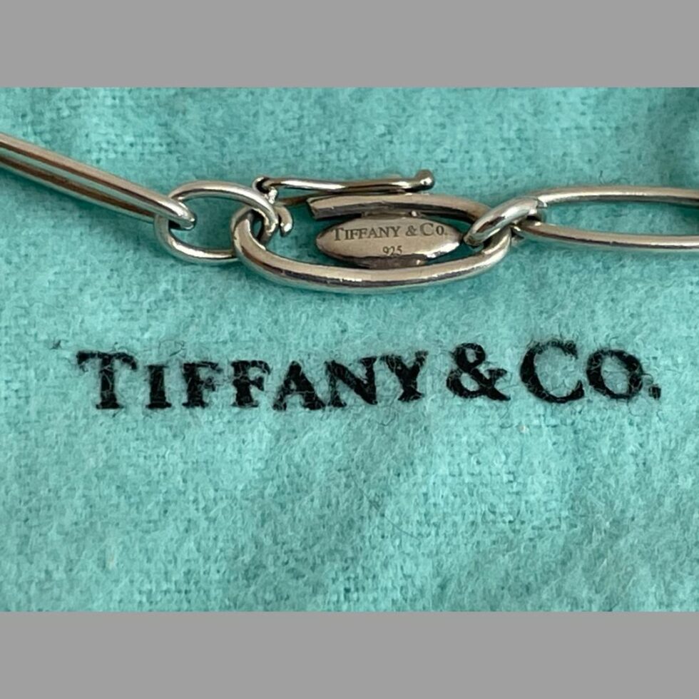 Close up photo the unusual clasp area of the bracelet which is still displayed on top of its pouch. The clasp is shown closed. It mimics the links of the bracelet in shape and has a hinged closure. The ring clasp on the end threads through the hinge when opened and hooks onto the clasp link. The hinge is then closed by popping the hook onto the small ball. Within this clasp link is a small plaque with hallmarks, makers marks and designers signature.