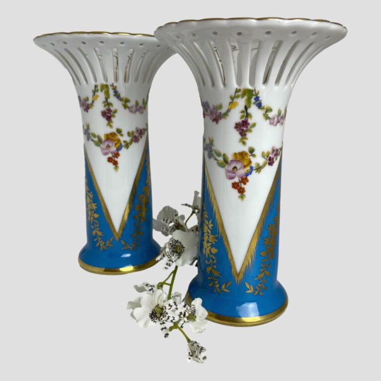 Main photo of Sèvres style porcelain vases displayed side by side with one slightly back from other. The photo is taken from a near eye level and there is a spray of white orchids in between the 2 vases. The main body of the vase is in white ending in a long V shape at the base where a vibrant dark turquoise blue sits. To the side, the white V becomes inverted to an upside down blue V. Where the white is seen most, there are 2 rows pretty multicoloured small flower garlands which continue all the way around the vase. To the top, the fluted tops have perforated sides like flower petals with a single dot perforation at top of each. Each perforated area is neatly finished in gilt. Such pretty vases - flowers not required!