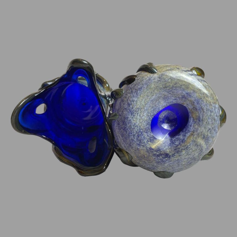 Photo of both signed studio glass vases laid on a flat surface side by side this time with the 2 pointed tip vase showing the base and looking into the 3 point tipped vase. The base of the 2 point tipped vase is mostly opalescent flecks with the centre only in blue. You can see the thickness of the black glass beading around the vase as it sticks out from the sides.