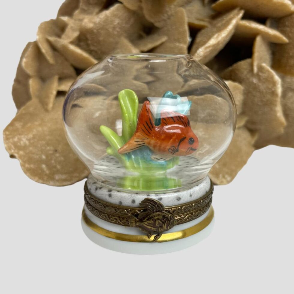 Main photo of Limoges goldfish bowl box displayed with the front of the box and goldfish in the foreground. The goldfish is an orangey red colour just like a goldfish and it has a smile on its face. Behind the fish is some seaweed in light blue and green. There is a rock of natural desert sand rose in the background for display only.