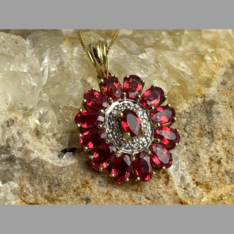 Main photo of ruby diamond pendant &chain displaying just the pendant forward facing and on top of a rock of natural river quartz. The light is shining on the stones which are lit up beautifully.
