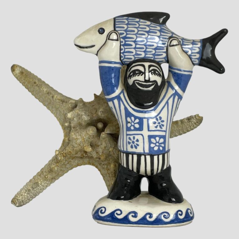 Main photo of fisherman by Bernard Moss seen with a starfish upright and behind for 'sea' effect. The fisherman is holding a large fish raised above his head with both arms up stretched. The whole studio art piece is in just 3 colours; black-hair, facial features, boots, fish tail and fin and face, blue-fish scales, decorations on jumper and waves and cream for everything else. This is such a smile inducing happy piece of studio pottery that anyone will love.