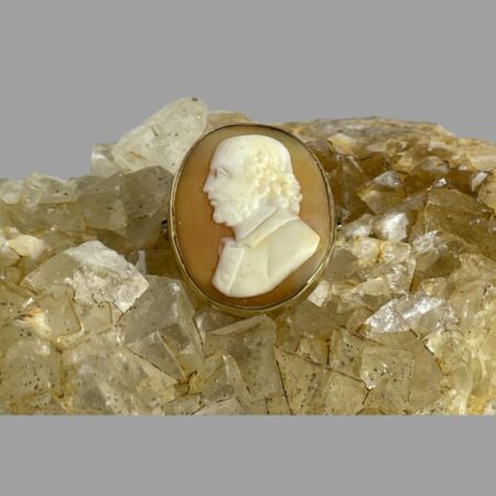 Main photo of 19th century Shakespeare cameo displayed upright on a rock of natural river quartz with the brooch front forward facing. The profile of William Shakespeare is left facing which is unusual for any cameo. He is wearing a wide collared top with his distinctive curly hair at neck length. Beautifully carved, a wonderful gift for a fan.