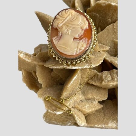 Main photo of 9Kt gold cameo brooch displayed perched on a rock of natural desert rose and shown with the brooch forward facing. The woman is in profile to the right and looking slightly downward with a pensive expression. Beautifully carved, her hair is up with a band adornment and she has a flower to her right shoulder. The outer rim of the brooch has 2 gold twist frames - the inner frame is a tight twist and the outer one a larger and wider twist. A very dreamy piece!