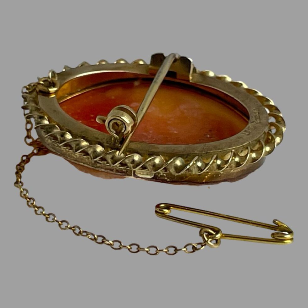 Photo of 18kt gold cameo brooch displayed facedown on a flat surface with the safety chain in the foreground in a sweeping curve. The clasp is undone and the pin sits just outside of the locking C clasp