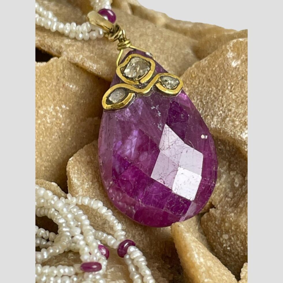 A close up photo of the ruby and diamond pendant. The light is shining on the ruby showing up the faceted detail to the generous sized stone. At the top of the teardrop shaped ruby is the largest of the 3 rose cut diamonds which has a frame of gold around it. Below this are the 2 smaller rose cut diamonds to the left and right. A fine gold rod is threaded through the top of the ruby and neatly twisted to attached to the gold bail. There are some minor scratch marks to the ruby.