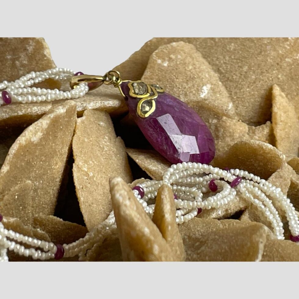 Photo of ruby diamond and pearly necklace still displayed on the desert sand rose rock this time with the ruby pendant front facing towards the top right corner to show the ruby depth. Some of the seed pearls and ruby beads are clearly visible in the foreground.