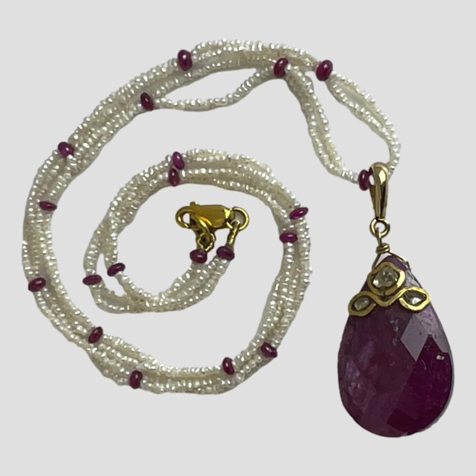 Photo of ruby diamond and pearl necklace displayed on a flat surface with the large ruby and diamond pendant in the right bottom corner and the seed pearl ruby bead necklace extended up and coiled in a swirl to its left.