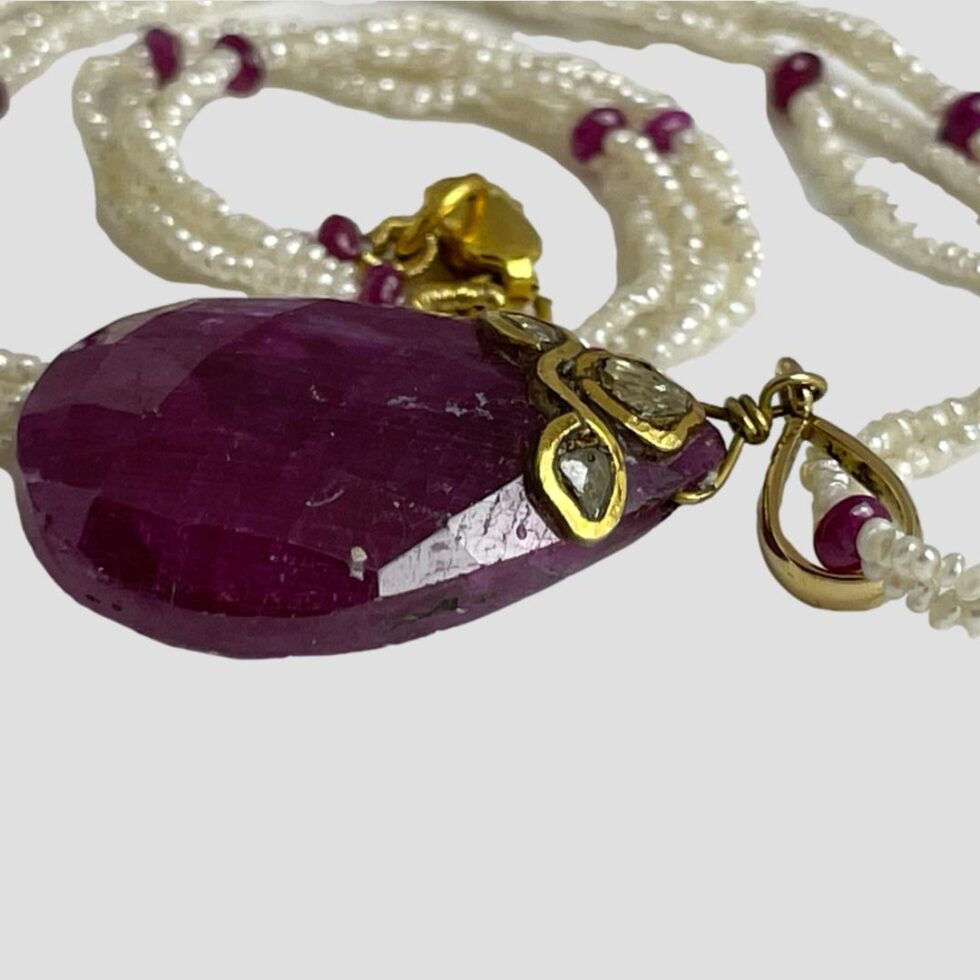 A close up of the gold areas of the necklace. The ruby pendant is laid flat with the wider end of teardrop on the left. The bail size of pendant is visible with the seed pearls and ruby beads going through. Looks as though it is possible to remove the pendant but we do not recommend it.