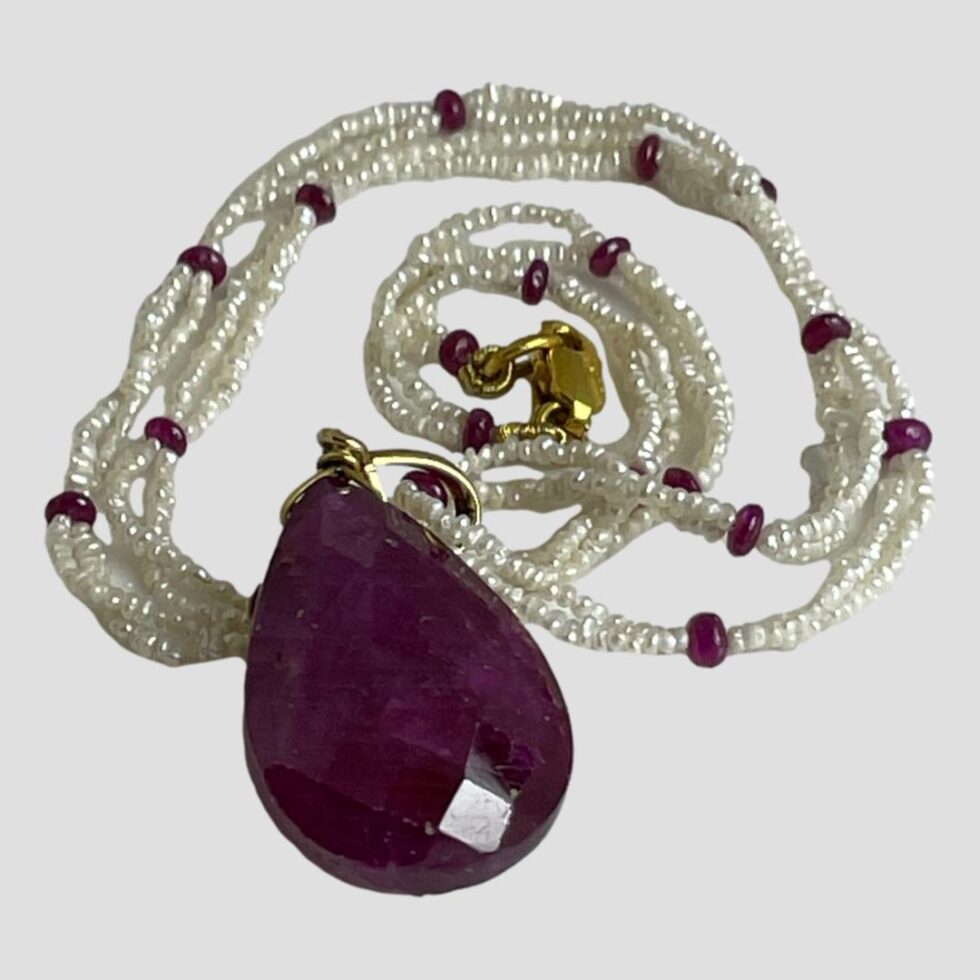 Photo of ruby diamond pearl necklace on a flat surface with necklace swirled round in a coil and pendant in the foreground displaying the back without the diamonds.