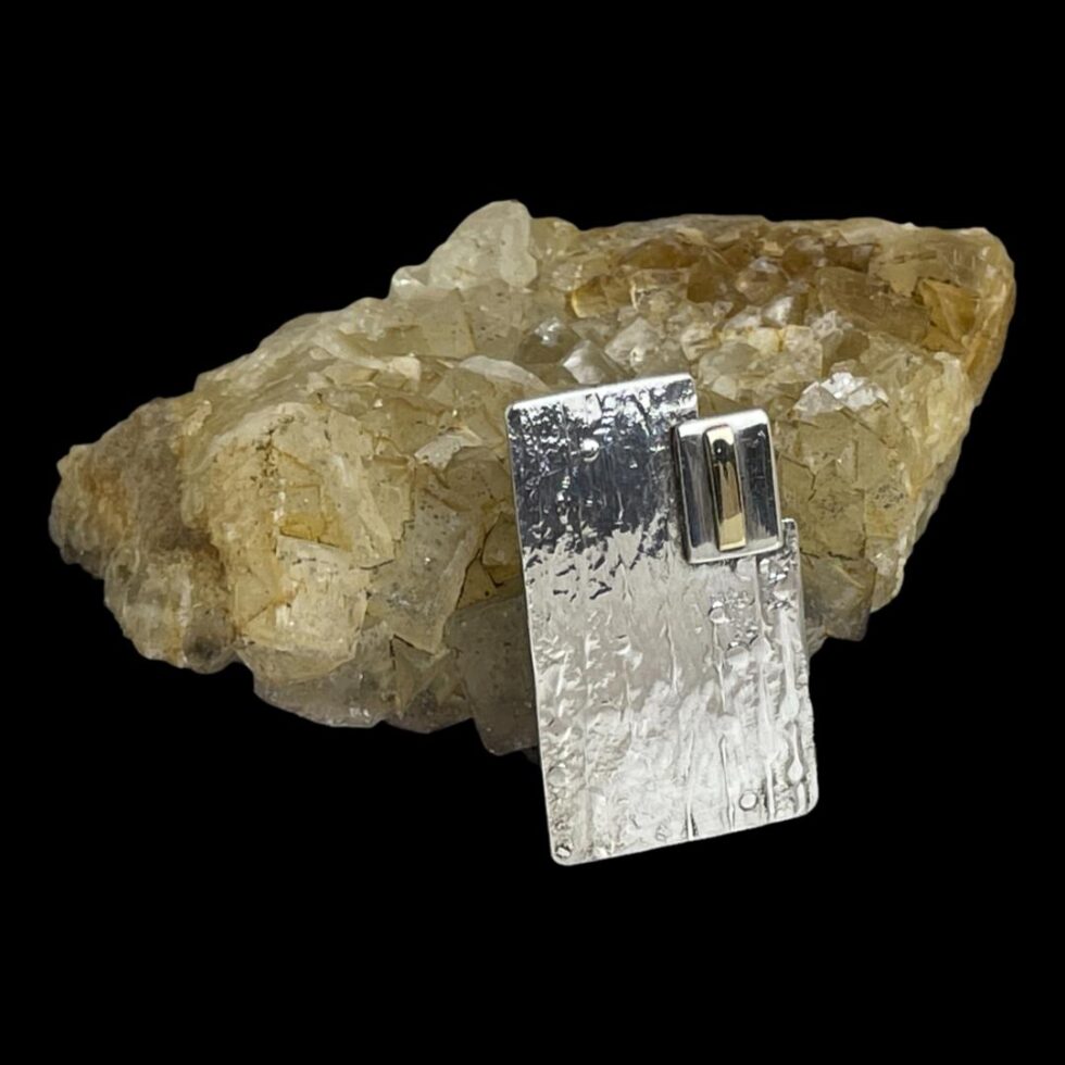 Photo of Modernist style silver brooch shown leaning against a rock of natural river quartz and the brooch front facing slightly towards the bottom right corner. The brooch is shown lengthways standing with the cut away area at the top right corner.