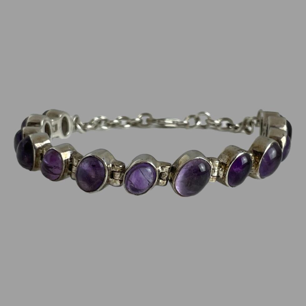 Photo of sterling silver amethyst bracelet displayed in a circle on a flat surface. The clasp area is closed & in the background. The bracelet, with its unique rigid setting of the links, is staying upright on its own accord and the leaning design of the amethyst cabochons is clearly evident here.