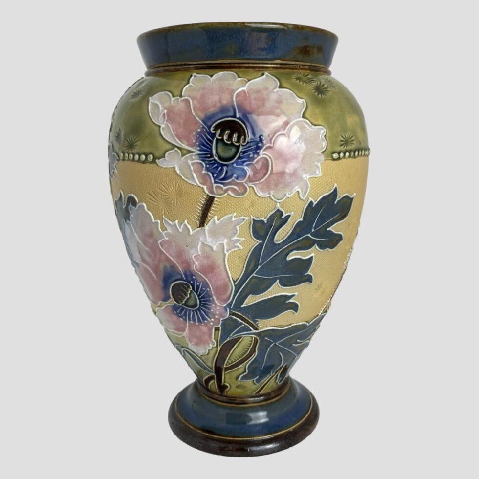 Main photo of Royal Doulton poppy vase seen from an eye level with 2 of the pink poppy blooms on display. The poppy at the top sits against the green at the top of the vase while the lower poppy remains mostly in the mute yellow middle area with just a petal of 2 invading the green around the bottom rim. The leaves match the dark teal blue around the lip and base with the blue of the poppy pistil a brighter shade.