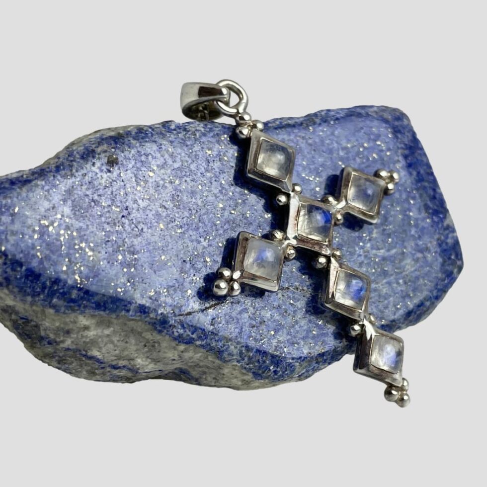 Photo of cross still displayed against a small rock of lapis lazuli with the front of cross facing to the right. 4 of the stones are now flashing the brilliant blue of the rainbow moonstone as it catches the light.