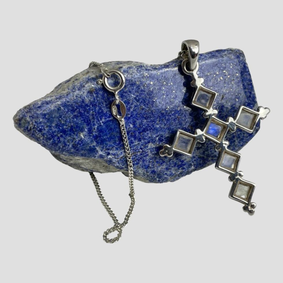 Photo of the back of silver moonstone cross &chain still leaning on a rock of lapis this time showing the back of pendant. The chain has been laid to the left of cross with the clasp area on show. The hallmark to the sealed pendant bail and the chain clasp is visible.