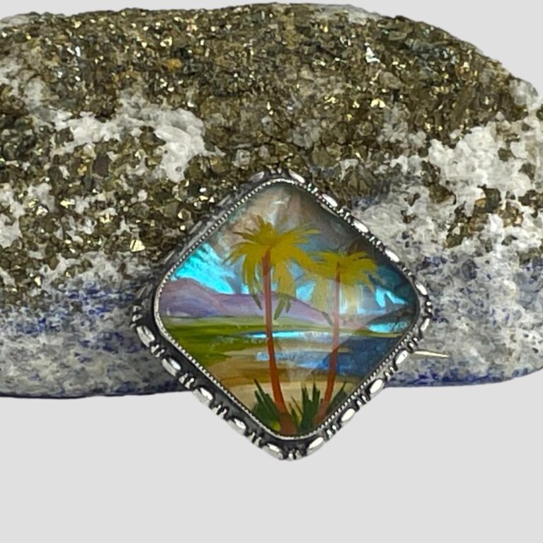 Main photo of silver butterfly wing brooch displayed leaning against a blue and gold flecked natural lapis rock with the brooch front forward facing. The brooch is to be worn in a diamond shape to match the image of tropical palms in the foreground and mountain range and lapping blue waves in the background.