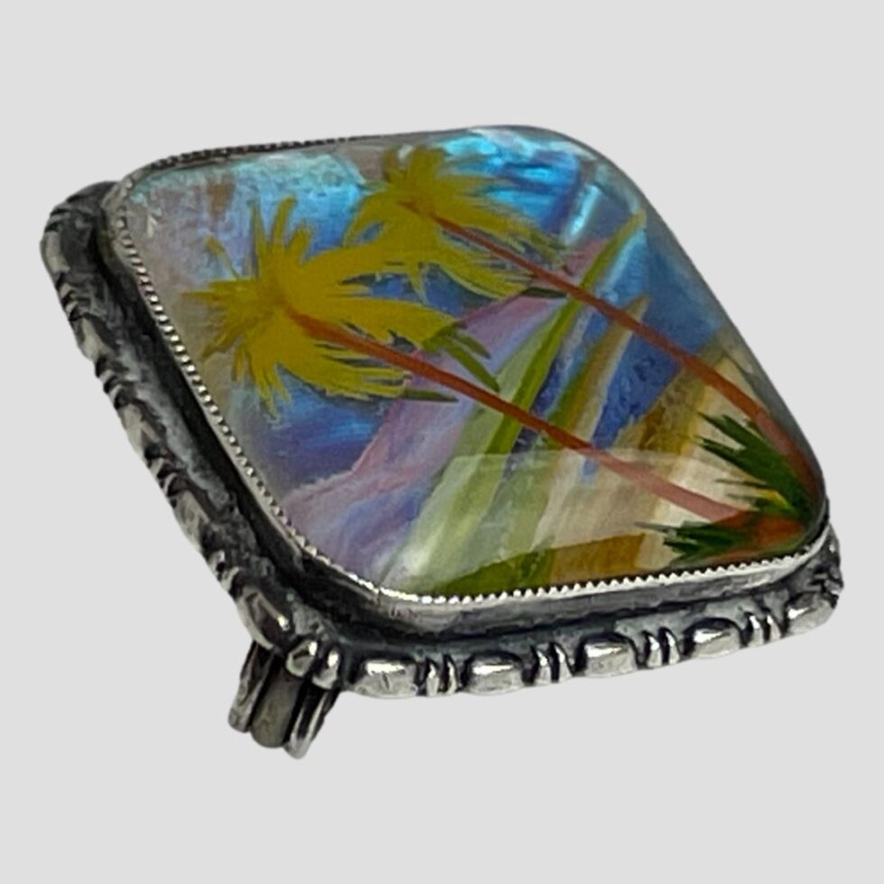 Photo of brooch displayed on a flat surface with the brooch front facing towards the top right corner of the photo. The lovely colours of the hand-painted scene with the blue butterfly wing additions is clearly visible here. The brooch also has a lovely frame all around to enhance the look.