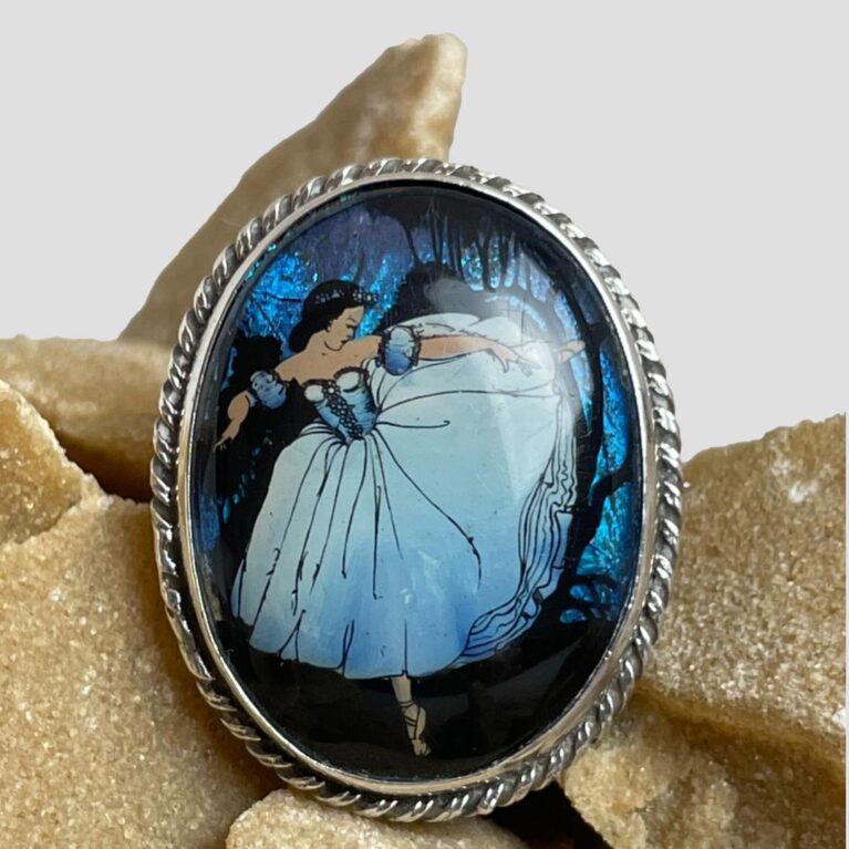 Main photo of Ballerina butterfly wing brooch displayed on top of a rock of natural desert sand rose and shown with the brooch front forward facing. The ballerina is painted in an arabesque pose and appears to be running away with the woods in the background. The woods, in electric blue, are all butterfly wings even the dark shadows of the trees. We are thinking Snow White.