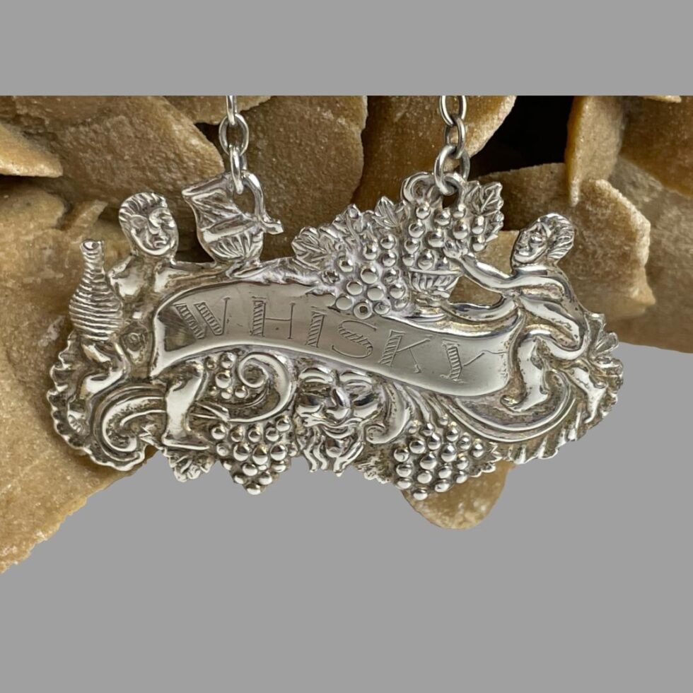 A close up photo of the silver whisky decanter label. At the centre is a wavy banner with WHISKY etched on. At the centre below the banner is an image of probably John Barleycorn - a traditional folk figure representing the agricultural cycle in the production of whisky. He is quite well camouflaged between grapes and grains to his left and right. At either end of the banner are 2 figures. On the left, carries a flagon with the jug attached to the chain end facing him as if filling the bottle. The one on the right appears to be lovingly tending to the barley or grapes which are sat in a bowl and attached to other end of the chain. Quite an elaborate label which gives more each time you look.