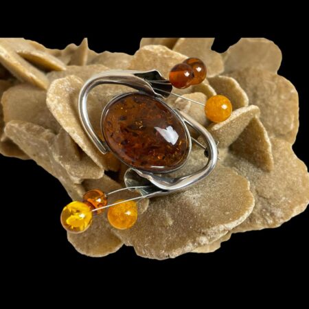 Main photo of sterling silver amber brooch displayed on top of a rock of natural desert sand rose. The central amber is a generously sized oval shape with the silver calla lily flower stems extending out from one side and the flower head leaning over the top of the large amber to finish on the opposite side where the stamen balls protrude. There is no top and bottom to this brooch so it can be worn any way round. The large central amber is a cognac colour with the stamen balls in cognac, butterscotch and honey colours. There is something quite space age looking about this lovely brooch and will definitely attract compliments! In this photo, the brooch is angled so the large oval amber is seen diagonally laid and the balls are positioned bottom left and top right corners.
