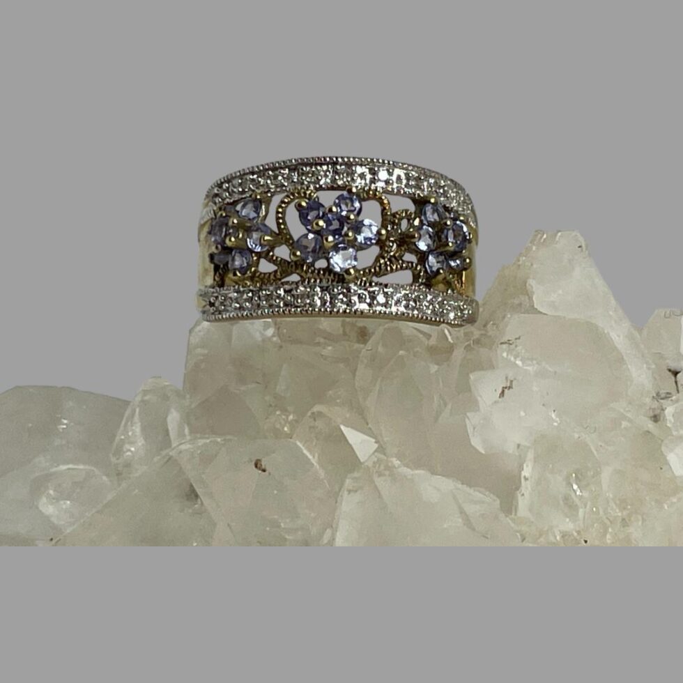 Main photo of modern gold tanzanite ring displayed on a rock of natural quartz crystal with the ring front forward facing. The ring has 3 flowers with 5 tanzanite petals and tanzanite centre to each horizontally in a line. Each flower head is separated with textured yellow gold vines. Like a border, the top and bottom edge of the ring front has a row of diamond chips which are set in white gold. The shoulders taper down to a thinner band at the back.