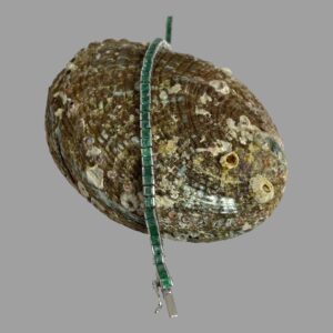Main photo of white gold emerald bracelet displayed laid across a seashell and shown vertically on the page. The square cut emeralds have an outer frame in white gold with no visible gold frame between the stones for the length of the bracelet. The green of the emerald is uniform throughout and vibrant in hue.