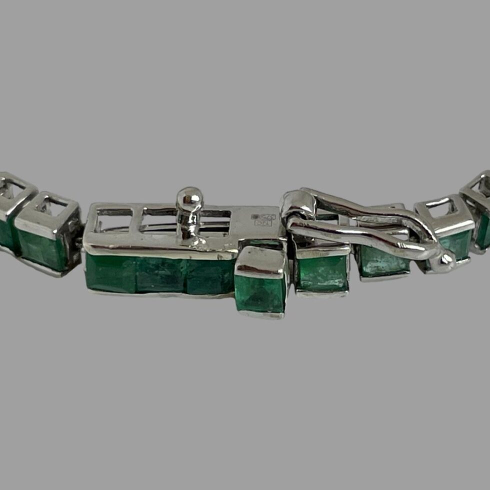 Close up photo of white gold emerald bracelet clasp shown pushed shut but with safety loop hook catch undone. The release button emerald raised from the other emeralds on the bracelet is well seen here.