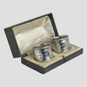 Main photo of the George V napkin rings displayed inside of their original fitted case. Some foxing and staining is visible to the silk on inside lid. The box is open to the bottom right.