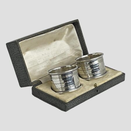 Main photo of the George V napkin rings displayed inside of their original fitted case. Some foxing and staining is visible to the silk on inside lid. The box is open to the bottom right.