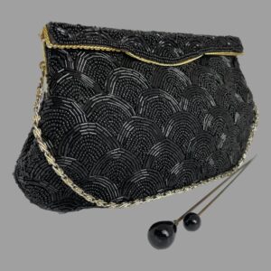 Main photo of vintage black beaded bag displayed set at a little angle with the front of bag facing towards the bottom right. The bag has a rigid opening flap which clips shut. There is a line of gold tone metal marking the flap opening. The bead pattern is uniform and bears resemblance to scales on a fish The chain handle is a silvery colour and is laid along the base of the bag. There are 2 black tipped hat pins in the foreground for effect.