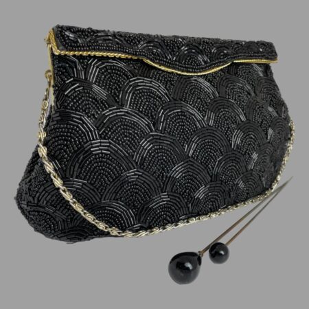 Main photo of vintage black beaded bag displayed set at a little angle with the front of bag facing towards the bottom right. The bag has a rigid opening flap which clips shut. There is a line of gold tone metal marking the flap opening. The bead pattern is uniform and bears resemblance to scales on a fish The chain handle is a silvery colour and is laid along the base of the bag. There are 2 black tipped hat pins in the foreground for effect.