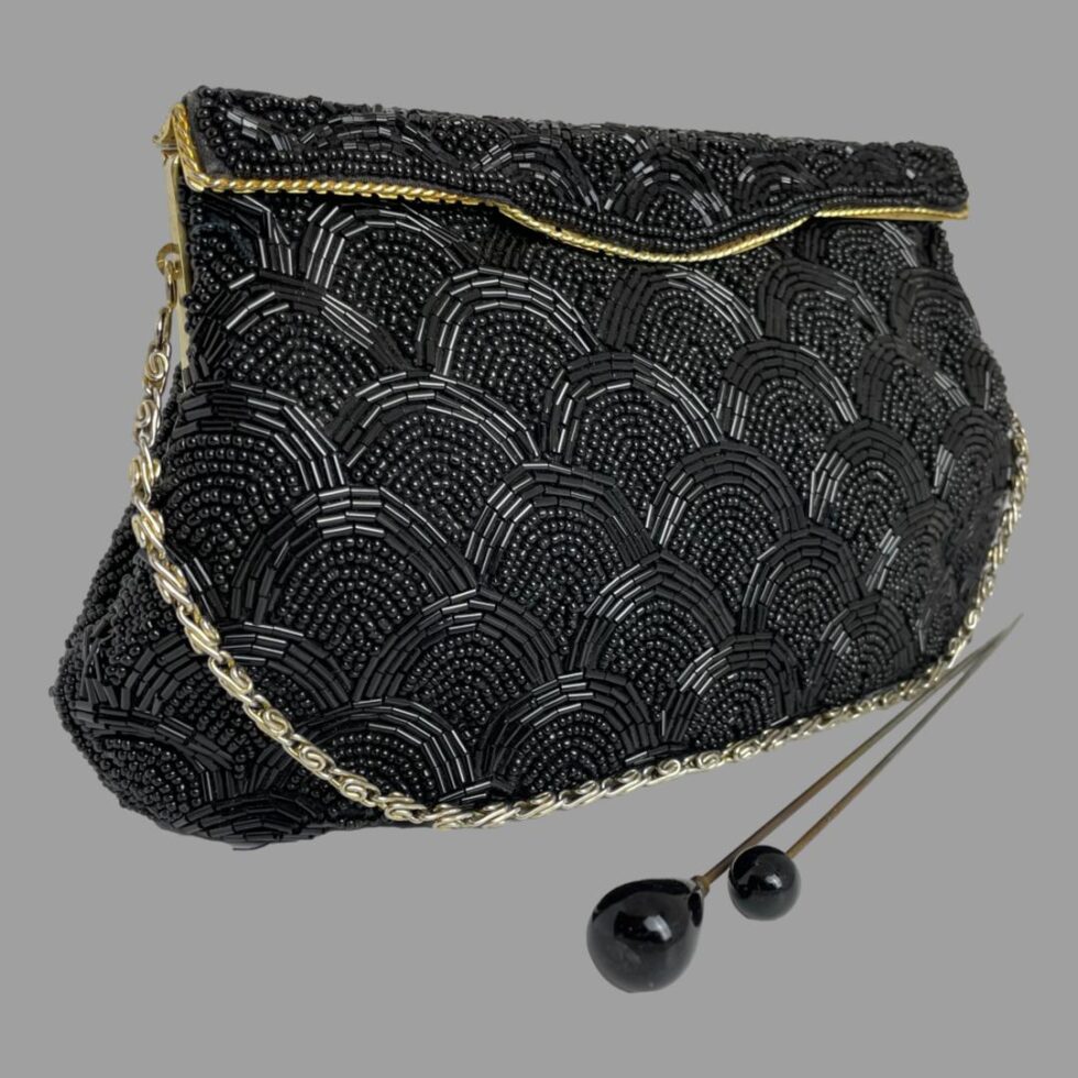Main photo of vintage black beaded bag displayed set at a little angle with the front of bag facing towards the bottom right. The bag has a rigid opening flap which clips shut. There is a line of gold tone metal marking the flap opening. The bead pattern is uniform and bears resemblance to scales on a fish The chain handle is a silvery colour and is laid along the base of the bag. There are 2 black tipped hat pins in the foreground for effect.
