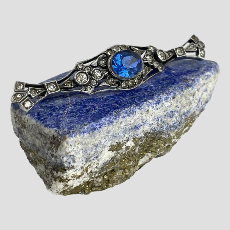 Main photo of c1900 French paste brooch displayed on top of a small rock of lapis lazuli with the brooch front facing towards the bottom right. The brooch has a bow like look about it with an ovular centre and fanned out ends to left and right. The blue stone in the centre is very vibrant and out blues the lapis. The clear paste stones are all present. A very pretty brooch.