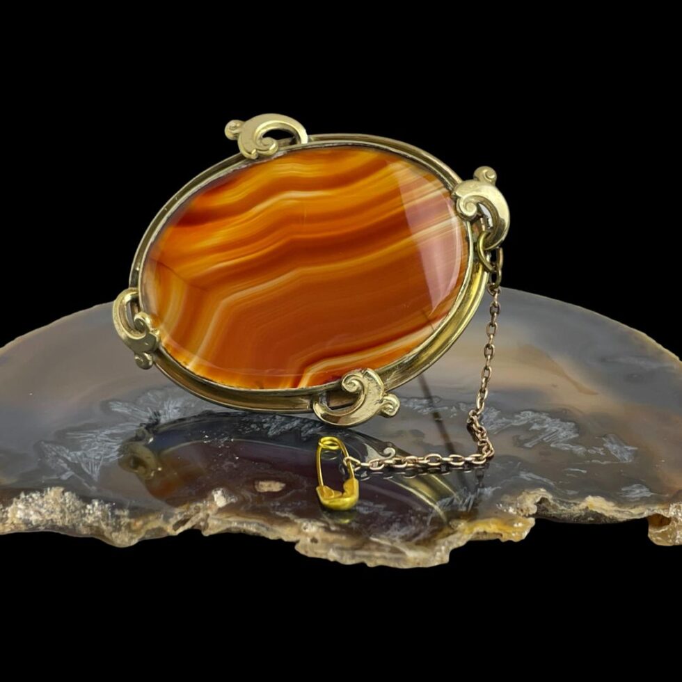 Main photo of Victorian banded agate brooch displayed on top of a slice of natural brown agate. The banded agate is a stunning deep terracotta red colour. The brooch has a pinchbeck frame with small wave shaped loops at 4 sides. On the wave loop on the right, the safety chain is attached and the pin has been placed in the foreground in front of the brooch.