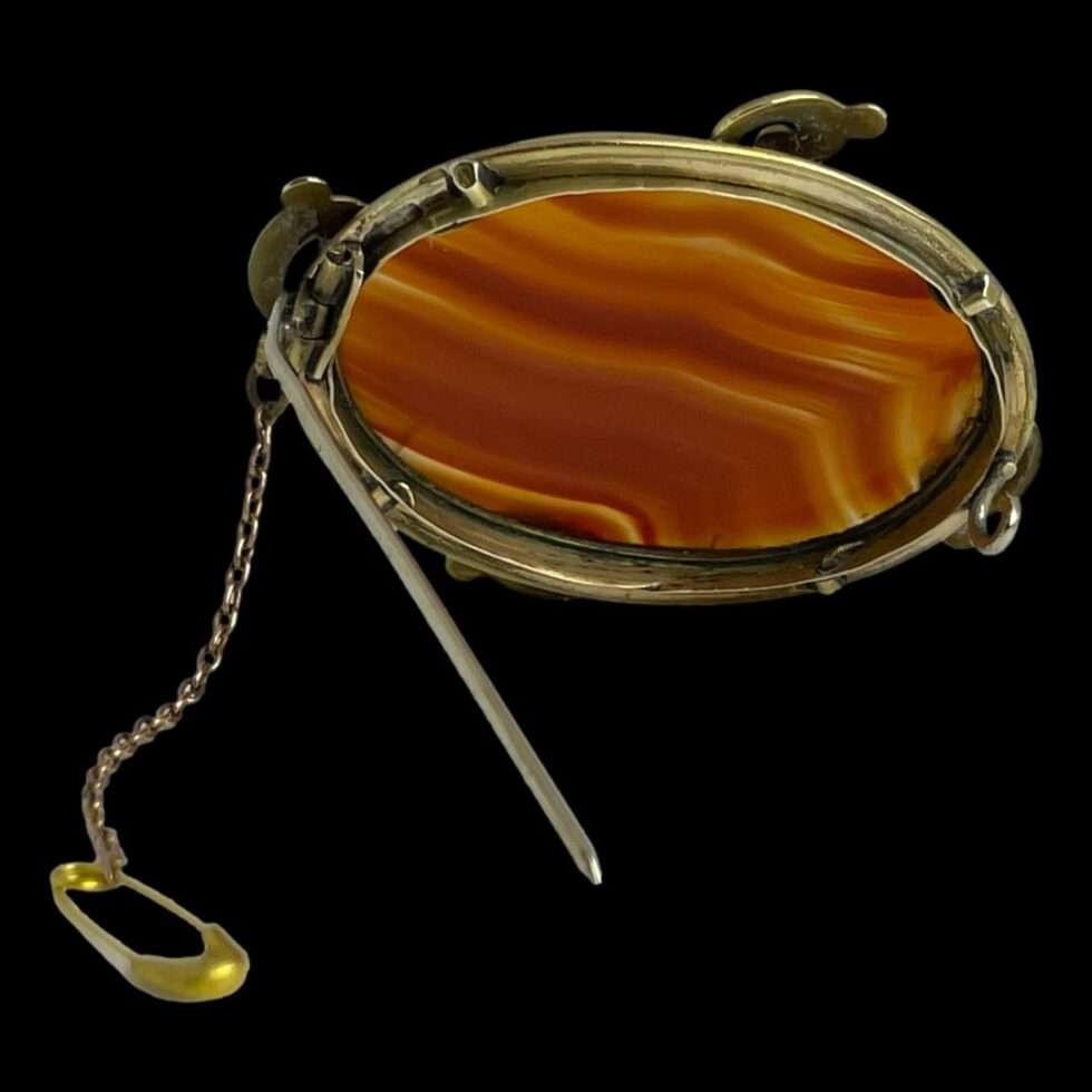 Photo of back of Victorian banded agate brooch seen on a black surface and displaying the brooch back. The brooch is propped upright by its own pin and the safety chain is extending out from the left.