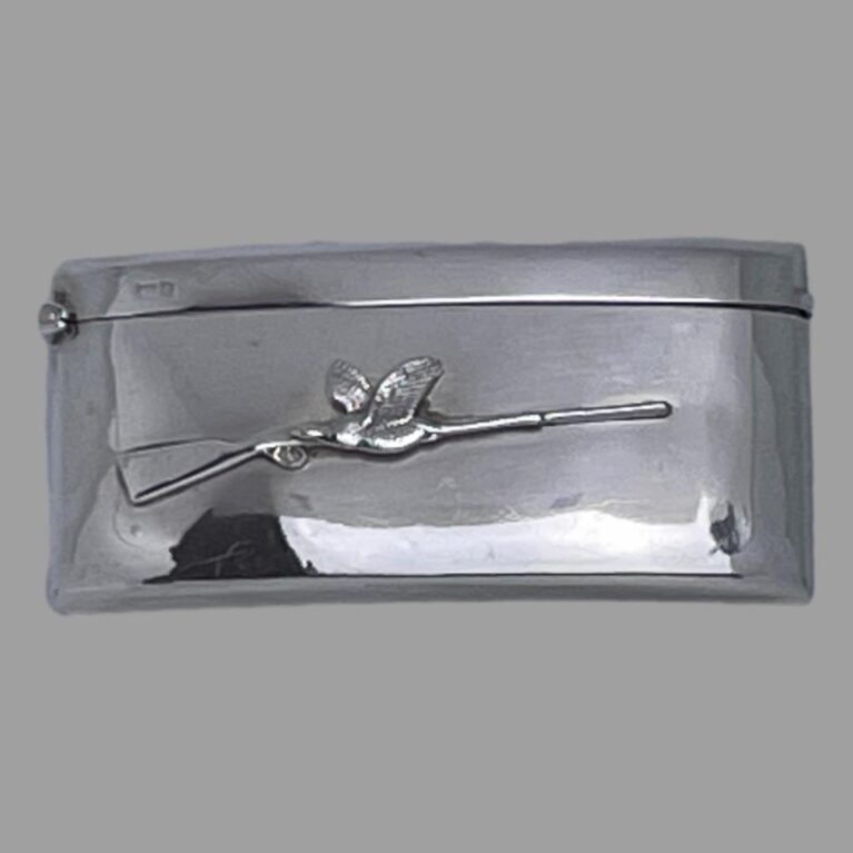 Main photo of antique business card case displayed on a flat surface and looking down at the front of the case. (The studio light and cover is reflecting on case in the shot) The case has a curved design so it will sit snuggly inside a back trouser or breast pocket. Other than the sleek design, the only decoration to this piece is a applied rifle at the centre with the stock end on the left and barrel on the right. In contrast, the single game bird is flying the other way passed the rifle and is situated at the centre of the rifle. This must be the one that got away thank goodness!