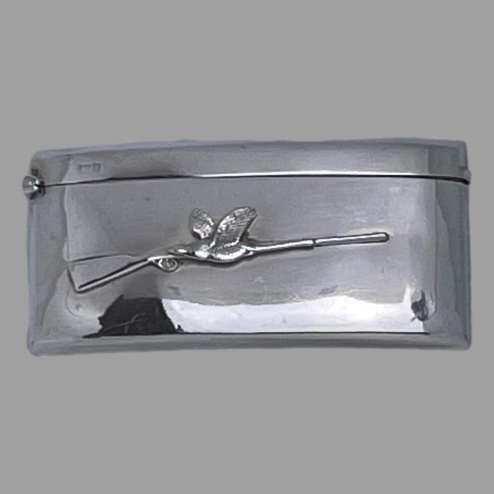 Main photo of antique business card case displayed on a flat surface and looking down at the front of the case. (The studio light and cover is reflecting on case in the shot) The case has a curved design so it will sit snuggly inside a back trouser or breast pocket. Other than the sleek design, the only decoration to this piece is a applied rifle at the centre with the stock end on the left and barrel on the right. In contrast, the single game bird is flying the other way passed the rifle and is situated at the centre of the rifle. This must be the one that got away thank goodness!