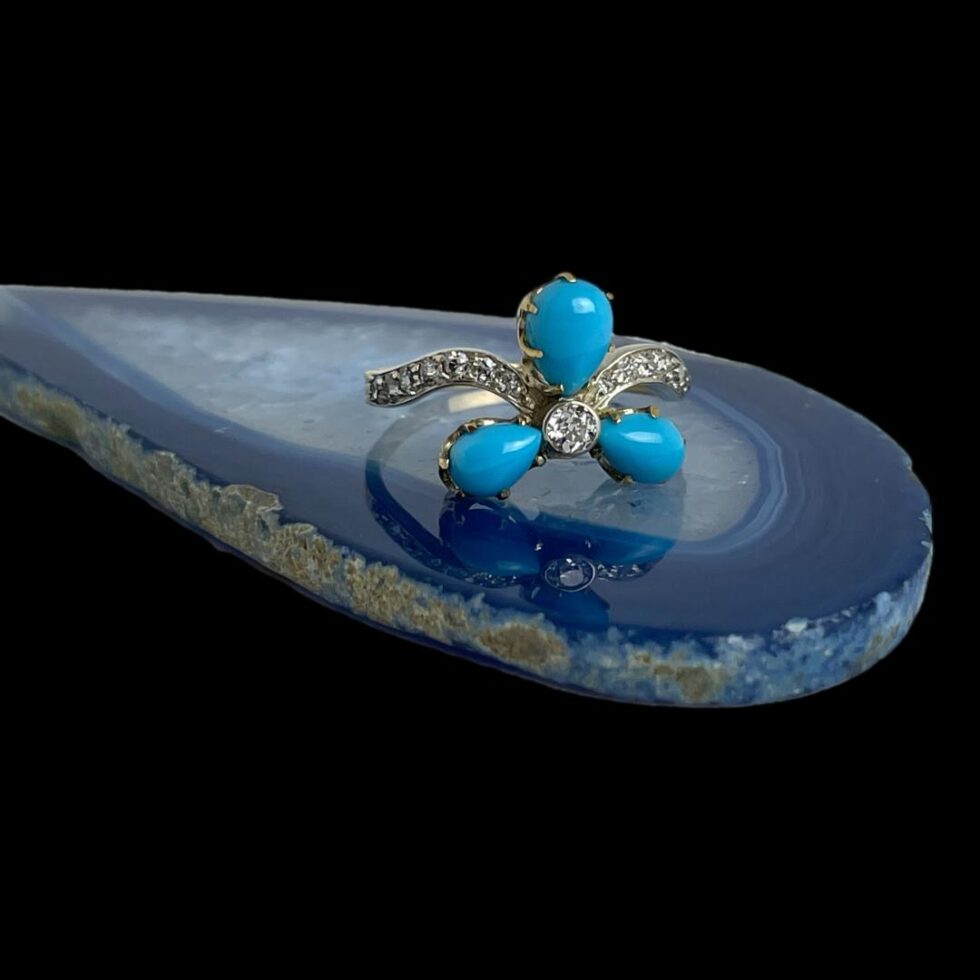 Main photo of diamond and turquoise ring displayed on top of a small slice of blue agate with the ring front forward facing. The petals consist of one large (at top in this photo) and 2 smaller (at lower left and right in this photo) teardrop shaped petals. The Victorian old cut diamond sits in the centre of the petals. 2 diamond encrusted bands curve out from the central diamond joining the shoulders which also have graduating diamonds. The ret of the band is in unmarked 15karat yellow gold. A very unusual stunning ring.