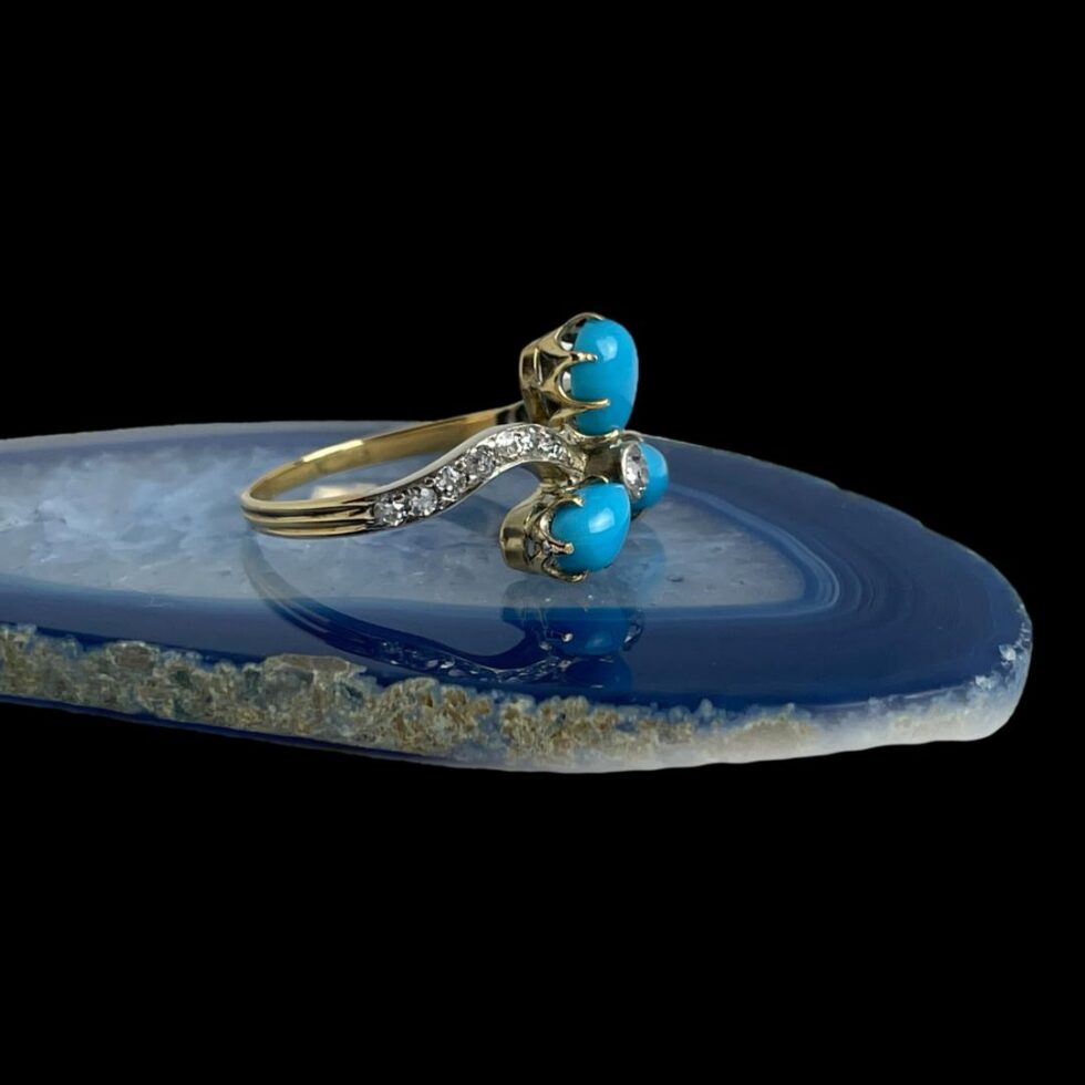 Photo of diamond and turquoise ring displayed on top of a slice of blue agate and shown with the ring front facing to the right showing the claw setting for the turquoise petals and one graduating diamond encrusted shoulder.