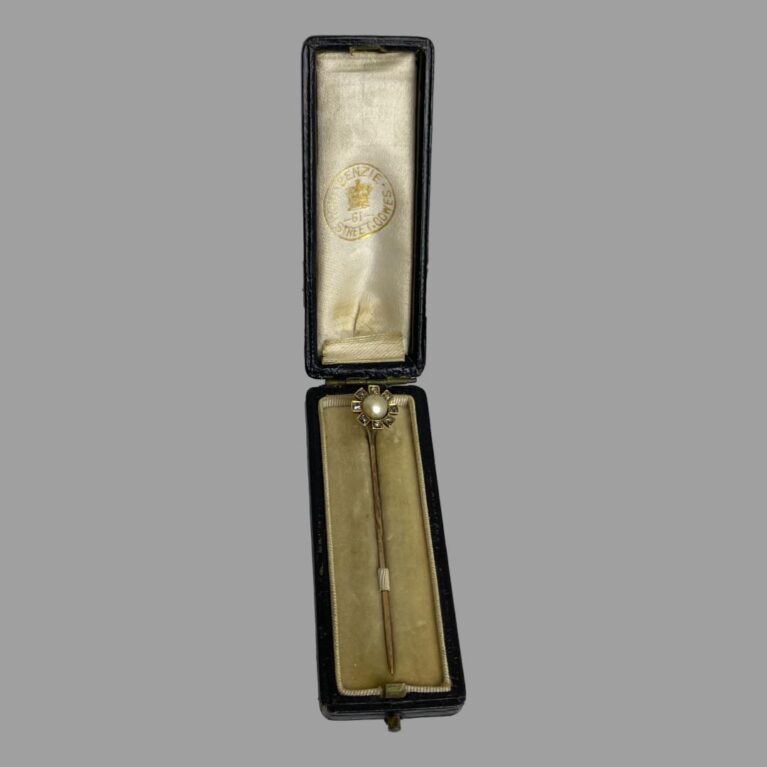 Main photo of Victorian stick pin displayed inside its original box. The pin is secured in place through a slot of cloth on the light mustard colour velvet interior. The silk on the lid is stamped with sellers mark in gilt for a shop in Cowes with year 61 - right in the middle of Queen Victorian's reign. The flowerhead of the pin has 8 gold 'petals' which have an old rose cut diamond to centre of each. The large pearl sits sunken in the centre.